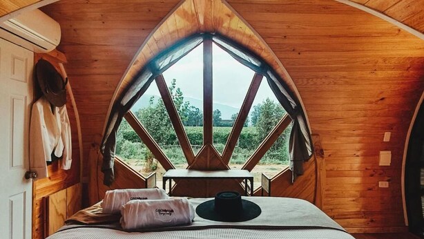 A cozy room with a bed, a table, and a view of the outdoors through a triangular window. - Colchagua Tiny Lodge (Santa Cruz)