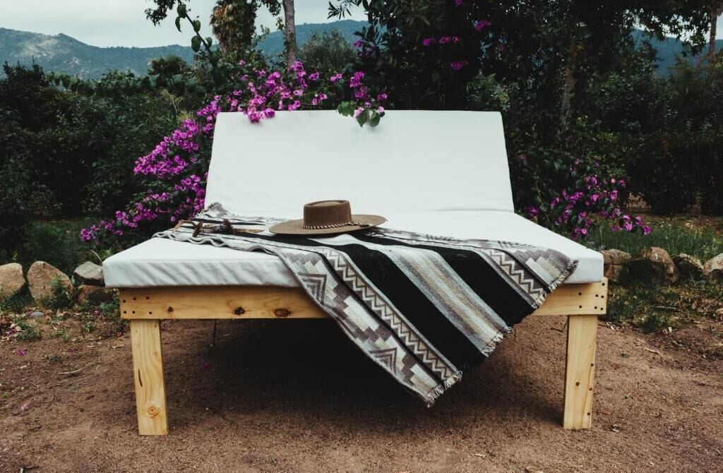 A wooden bed with a white mattress and a patterned blanket, set against a backdrop of purple flowers and greenery.