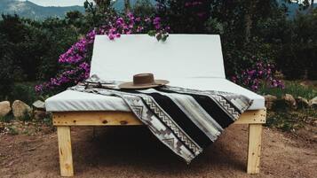 A wooden bed with a white mattress and a patterned blanket, set against a backdrop of purple flowers and greenery.