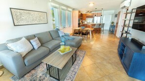 A modern living room with a grey sofa, a wooden coffee table, and a kitchen in the background. - VILLA CUQUI – Stunning Ocean Views & Poolside Paradise at Sandy Beach, Rincon (Rincon)