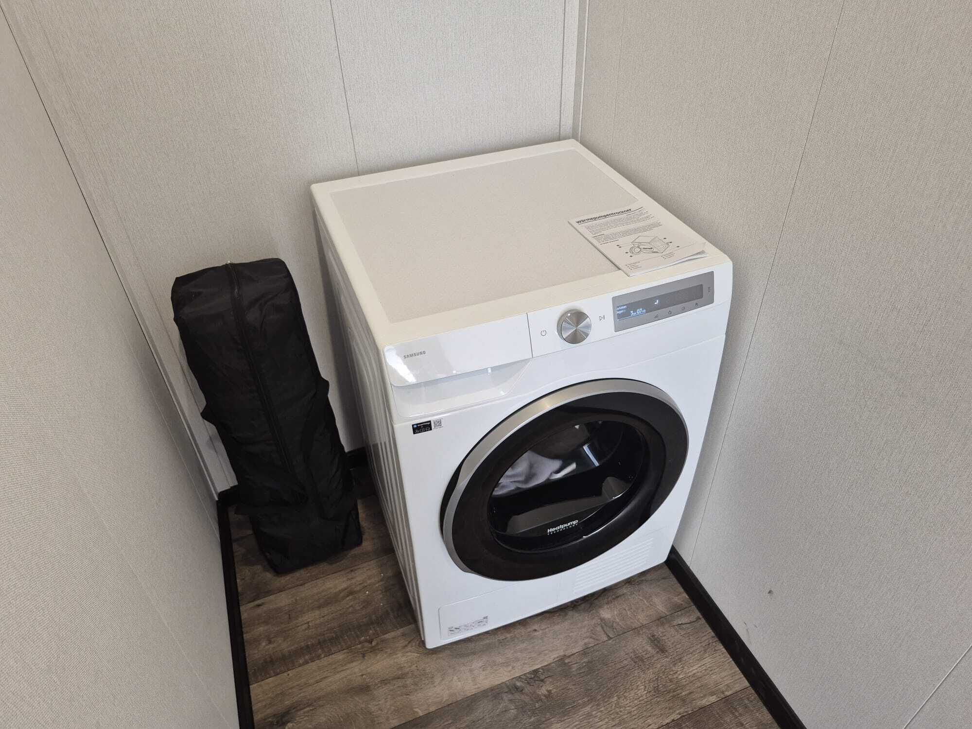 A compact, white front-loading washing machine in a corner of a room with wooden flooring and light-colored walls.