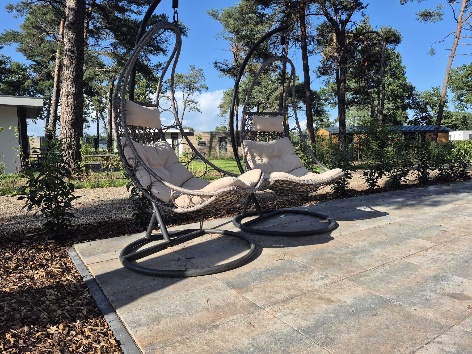Dos sillones de mimbre con cojines, ubicados en un patio de piedra rodeado de árboles.