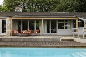 Un espace aménagé au bord de la piscine, comprenant un coin salon couvert, une piscine et un bâtiment en pierre doté d’une cheminée.