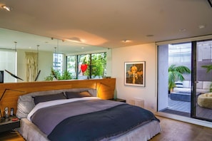 Un dormitorio moderno con una cama grande, una mesita de noche con lámpara, un espejo y un ventanal corredero que da a un espacio exterior con plantas.