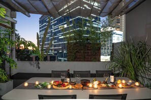Un área de comedor al aire libre con una mesa dispuesta para una comida, rodeada de plantas y velas, con vista a un edificio moderno.