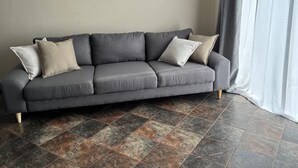 Un sofá gris con cuatro cojines en una habitación con suelo de baldosas y una cortina blanca.