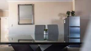 Un comedor moderno con una mesa de vidrio, dos sillas grises tapizadas, una pequeña planta en maceta, un espejo montado en la pared y un refrigerador empotrado.