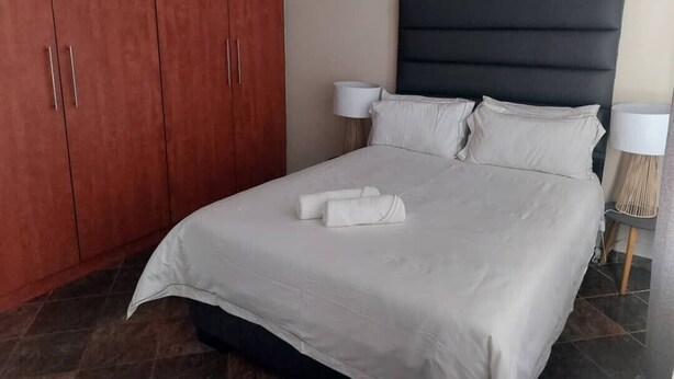 Una cama bien tendida con sábanas blancas y dos toallas enrolladas, un cabecero oscuro y dos lámparas de noche sobre un suelo de baldosas.