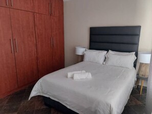 A neatly made bed with white linens and two rolled towels, a dark headboard, and two bedside lamps on a tiled floor. - Your holiday home, in the most quiet and yet secure neighborhood.    (Swakopmund)