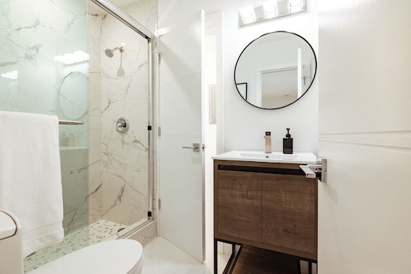 A bathroom with a glass-enclosed shower, a toilet, a sink with a wooden cabinet, and a round mirror on the wall. - • Palm Pointe • 0.3 Miles to beach - Heated pool (Fort Lauderdale)