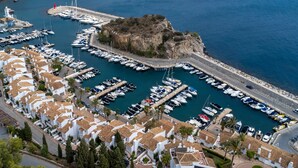 Uma marina com vários barcos atracados, um afloramento rochoso e edifícios brancos com telhados de terracota.