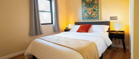 Una cama bien hecha con colcha blanca y beige, un cojín rojo y una mesita de noche con lámpara. Un cuadro floral adorna la pared sobre la cama.