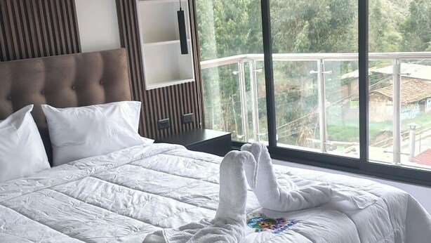 A large bed with white bedding and a towel, a wooden headboard, and a view of greenery through large windows. - HOTEL EL CASTOR UCHUYACU HUARAZ (Tarica)