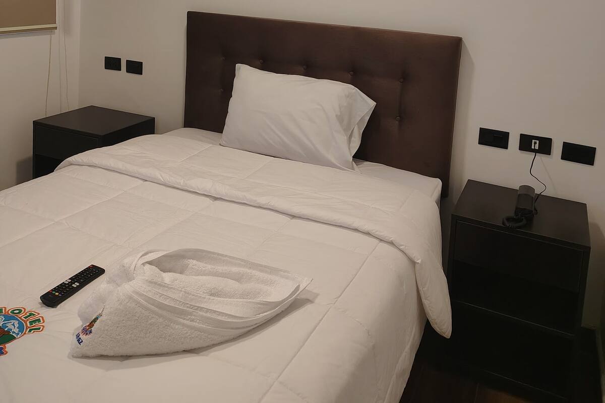 A hotel room with a bed, a nightstand, a remote control, and a towel on the bed.