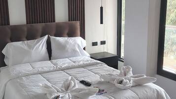 Una habitación de hotel moderna con una cama grande, vigas de madera en el techo y vistas a la vegetación. Se observa una mesita de noche con una lámpara.