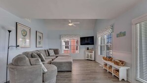 A living room with a sectional sofa, a ceiling fan, a television, and a window with blinds.