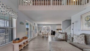 A spacious living area with a high ceiling, a staircase, and a dining area in the background.