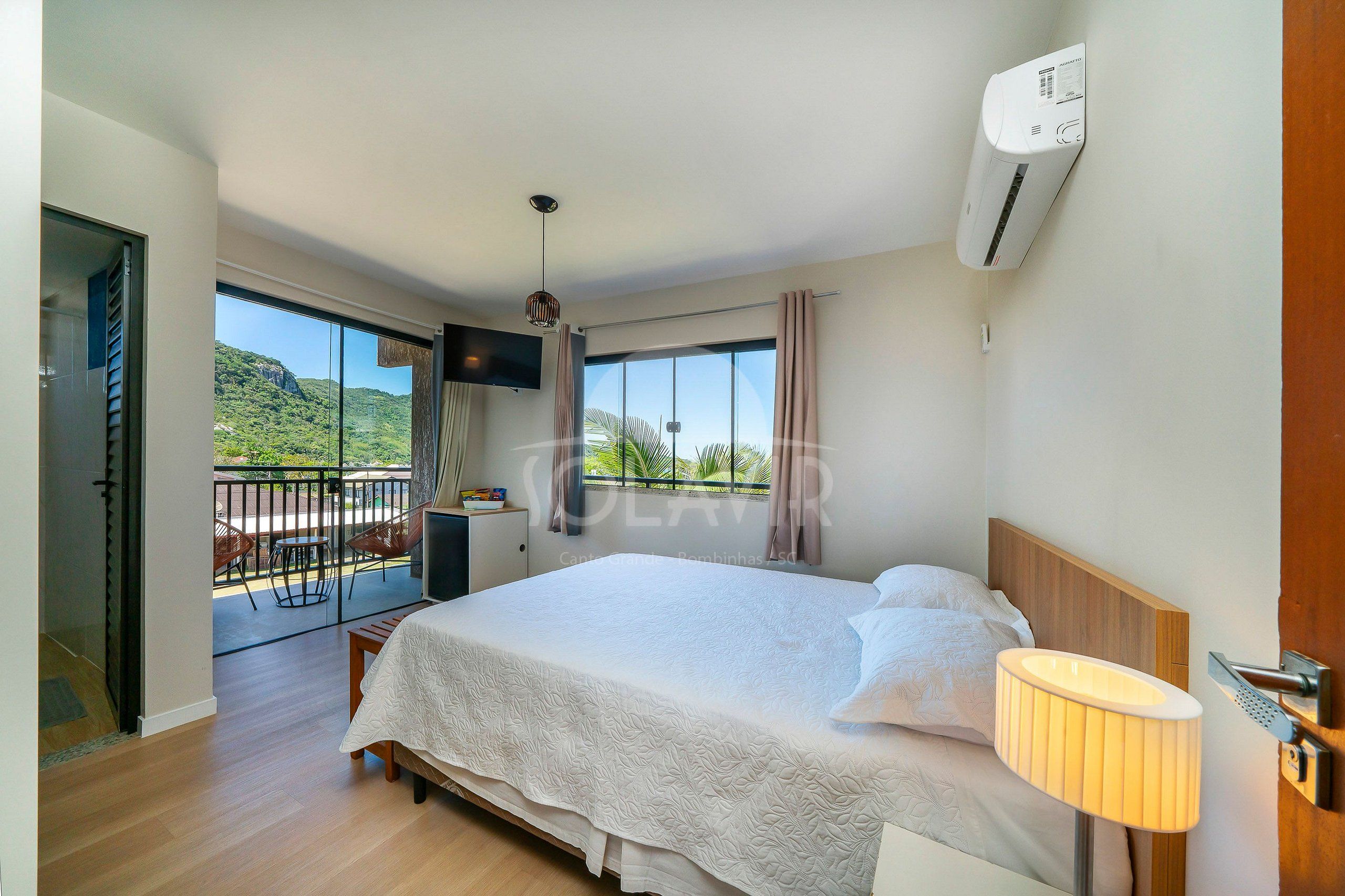 A hotel room with a bed, a bedside lamp, a TV, and a balcony with a view of greenery.