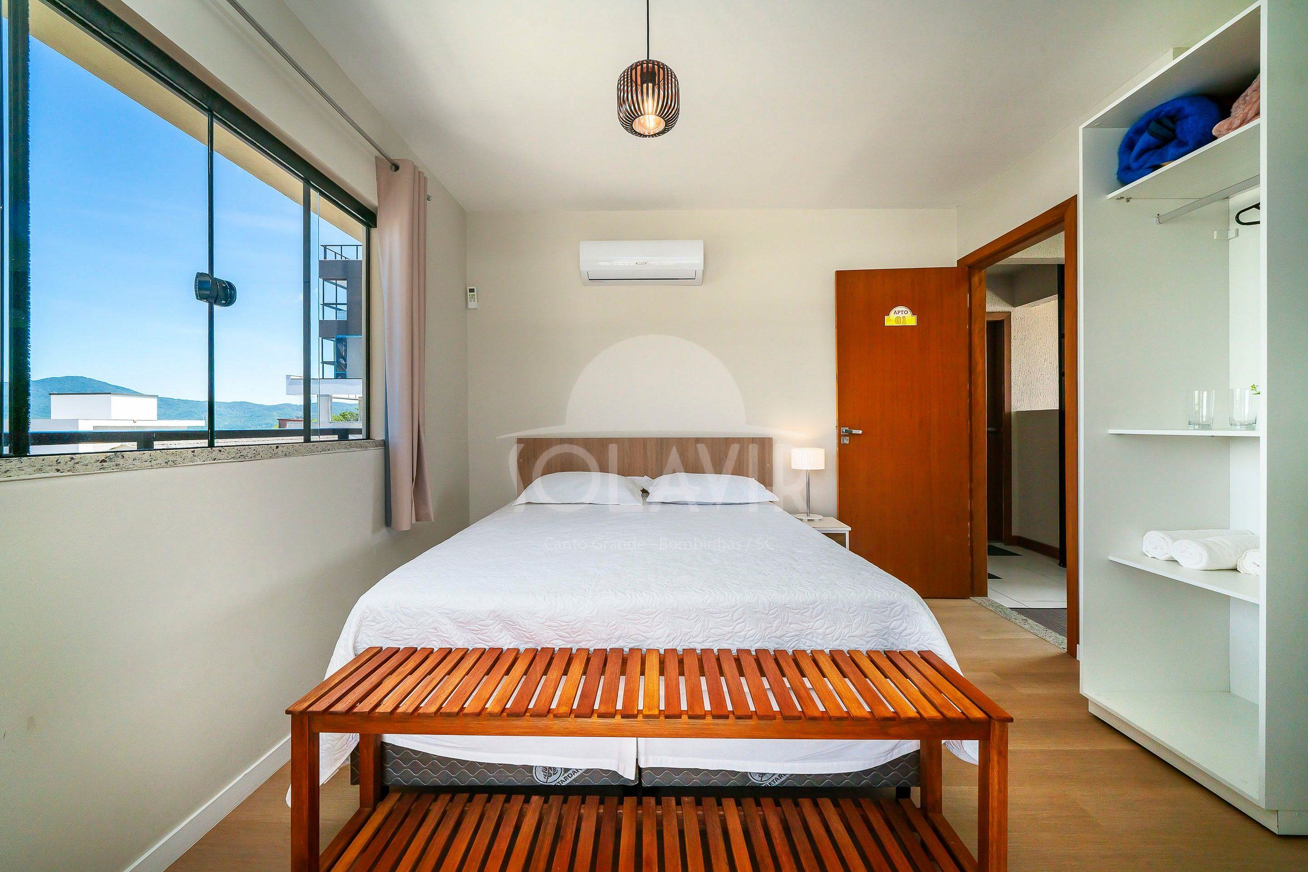 Un dormitorio con cama, un banco de madera, un armario y una ventana con vistas a edificios.