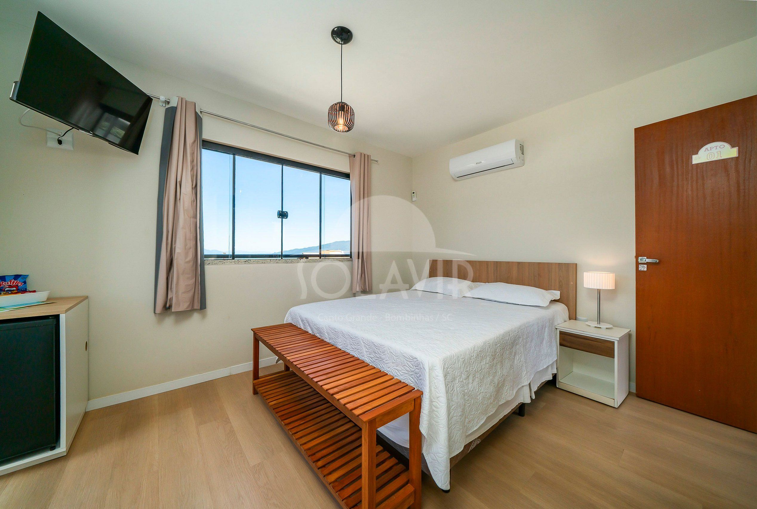A hotel room with a bed, bedside table, lamp, TV mounted on the wall, and a window with curtains.