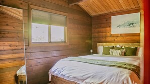 Een slaapkamer in een houten hutje met een bed, een raam en een schilderij aan de muur.