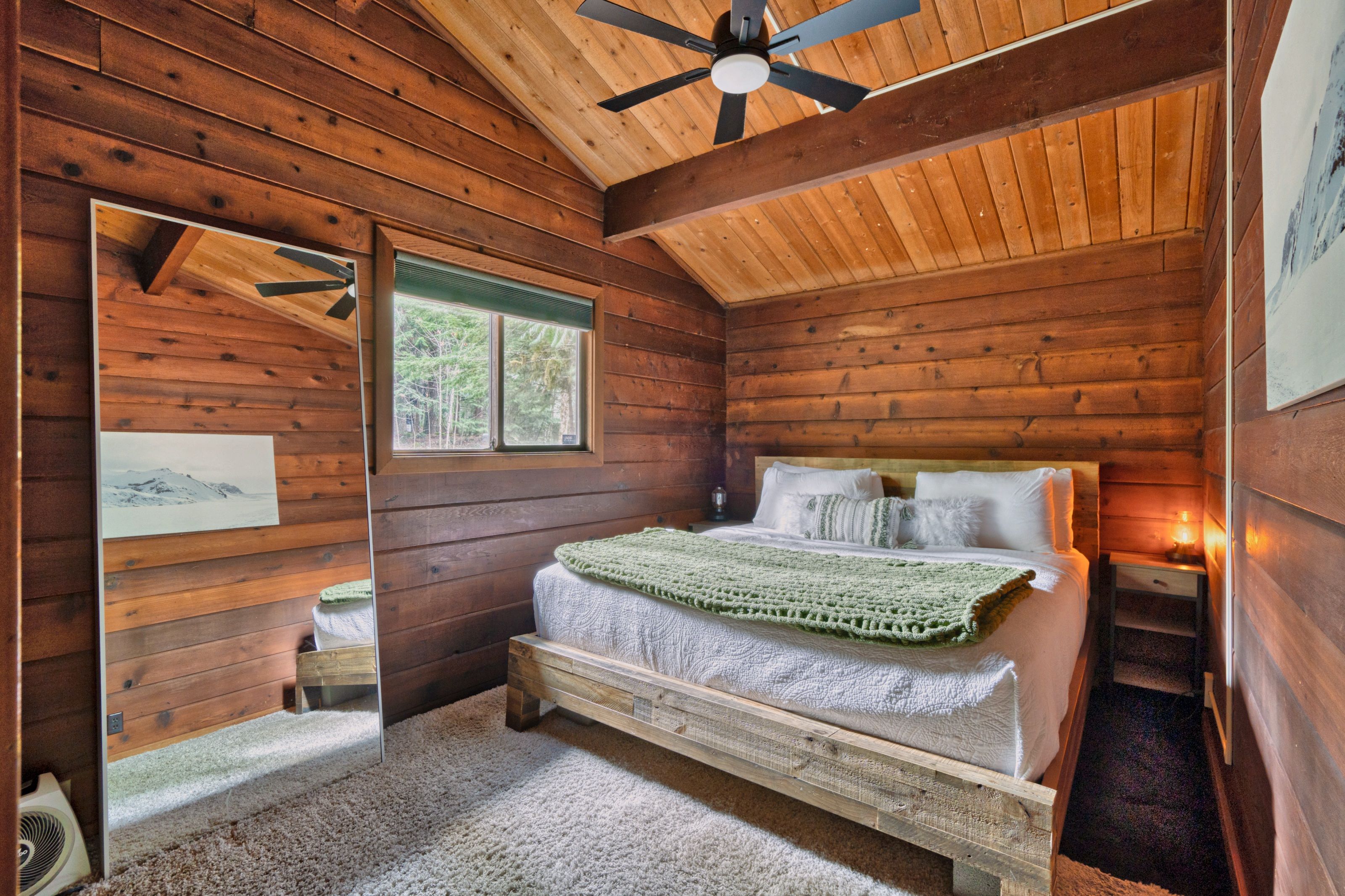 Een slaapkamer in een houten chalet, met een bed, plafondventilator, raam en spiegel.
