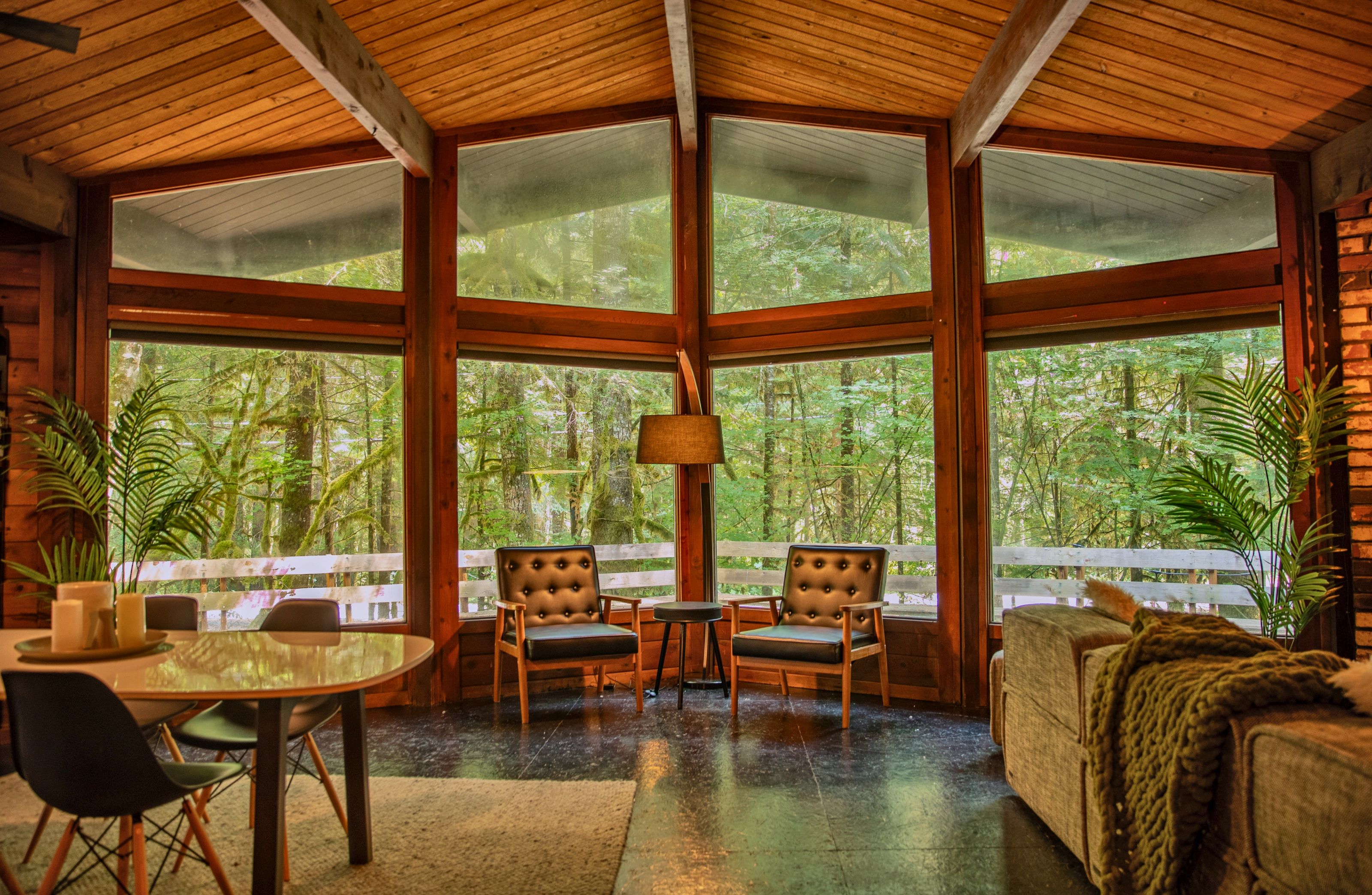 A room with large windows, a dining table, two chairs, a sofa, and a lamp.