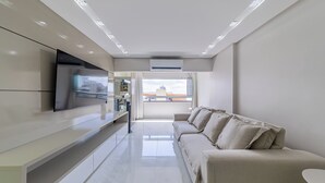 A modern living room with a sofa, TV, and a view of the city through large windows. - PASÁRGADA PONTA VERDE, WONDERFUL APARTMENT, CLOSE TO THE BEACH AND WITH SEA VIEW. (Ponta Verde Maceió)