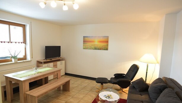 A living room with a wooden table, bench, black chairs, a red rug, a sofa, a TV, and a painting of a sunset.