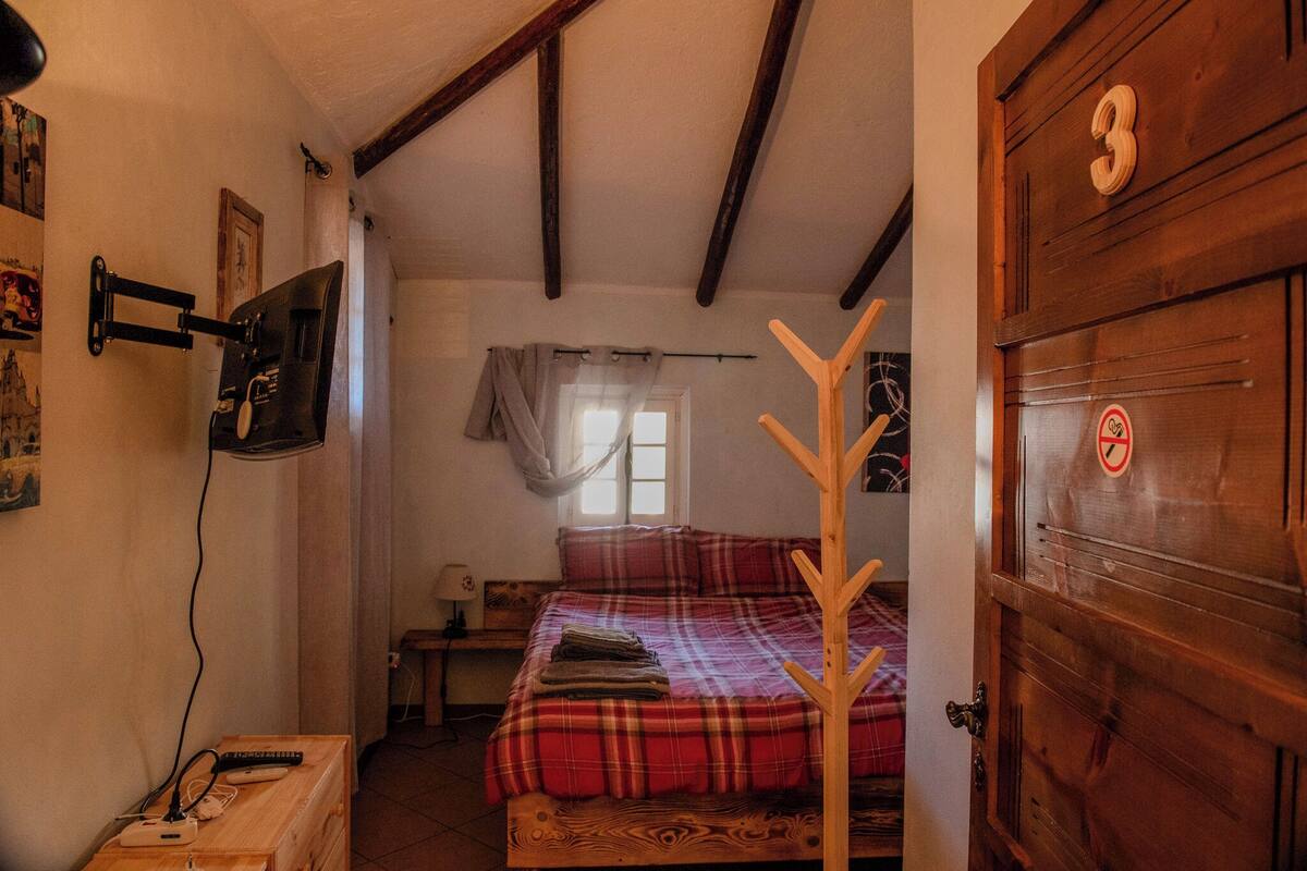 Un dormitorio con una puerta de madera identificada con el número 3, una cama con colcha de cuadros, un perchero y un televisor instalado en la pared.