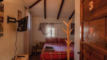 A bedroom with a wooden door marked with the number 3, a bed with a plaid bedspread, a coat rack, and a television mounted on the wall.