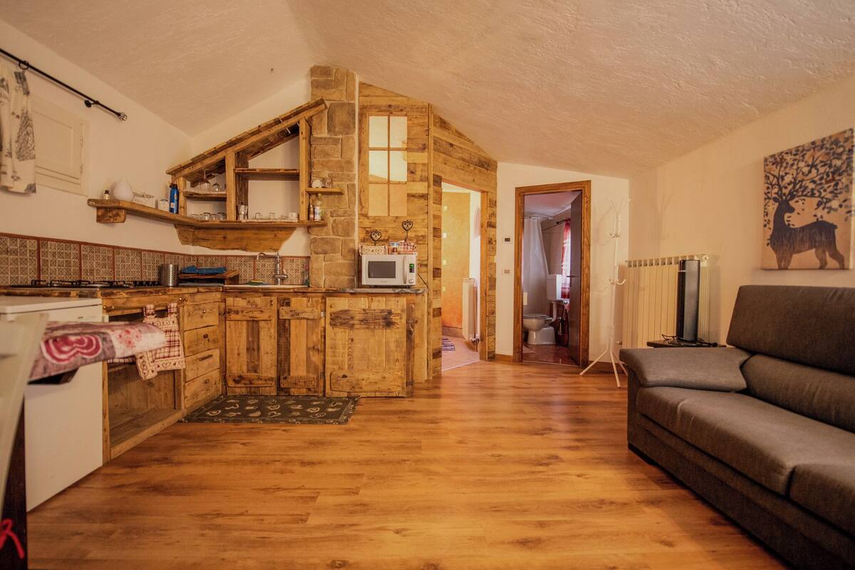 Una cocina rústica con armarios de madera y una pared de piedra, que da paso a una sala de estar con sofá y radiador.