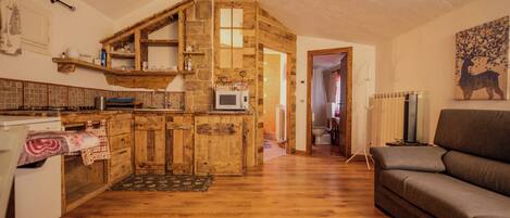 A rustic kitchen with wooden cabinets and a stone wall, leading to a living area with a sofa and a radiator.