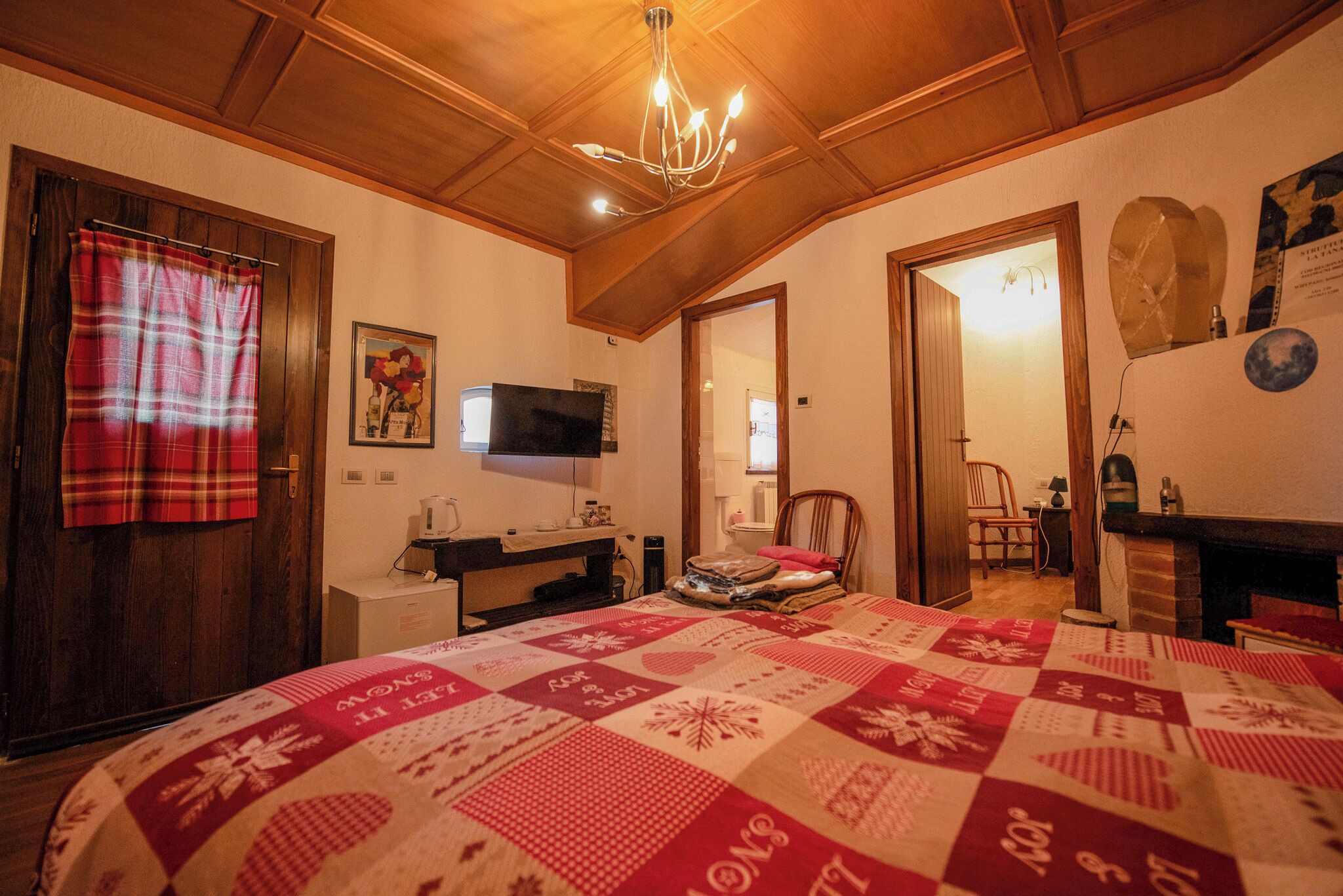 Ein gemütliches Schlafzimmer mit Holzbalkendecke, einem Bett mit rotem und weißem Quilt, einem an der Wand befestigten Fernseher und einem Kamin.