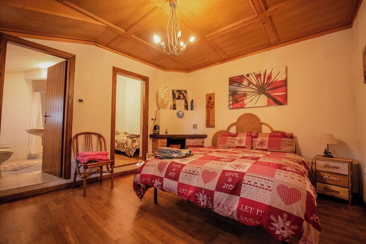 Un dormitorio con techo de madera, una cama con colcha roja y blanca, una mesita de noche, una silla y un espejo.
