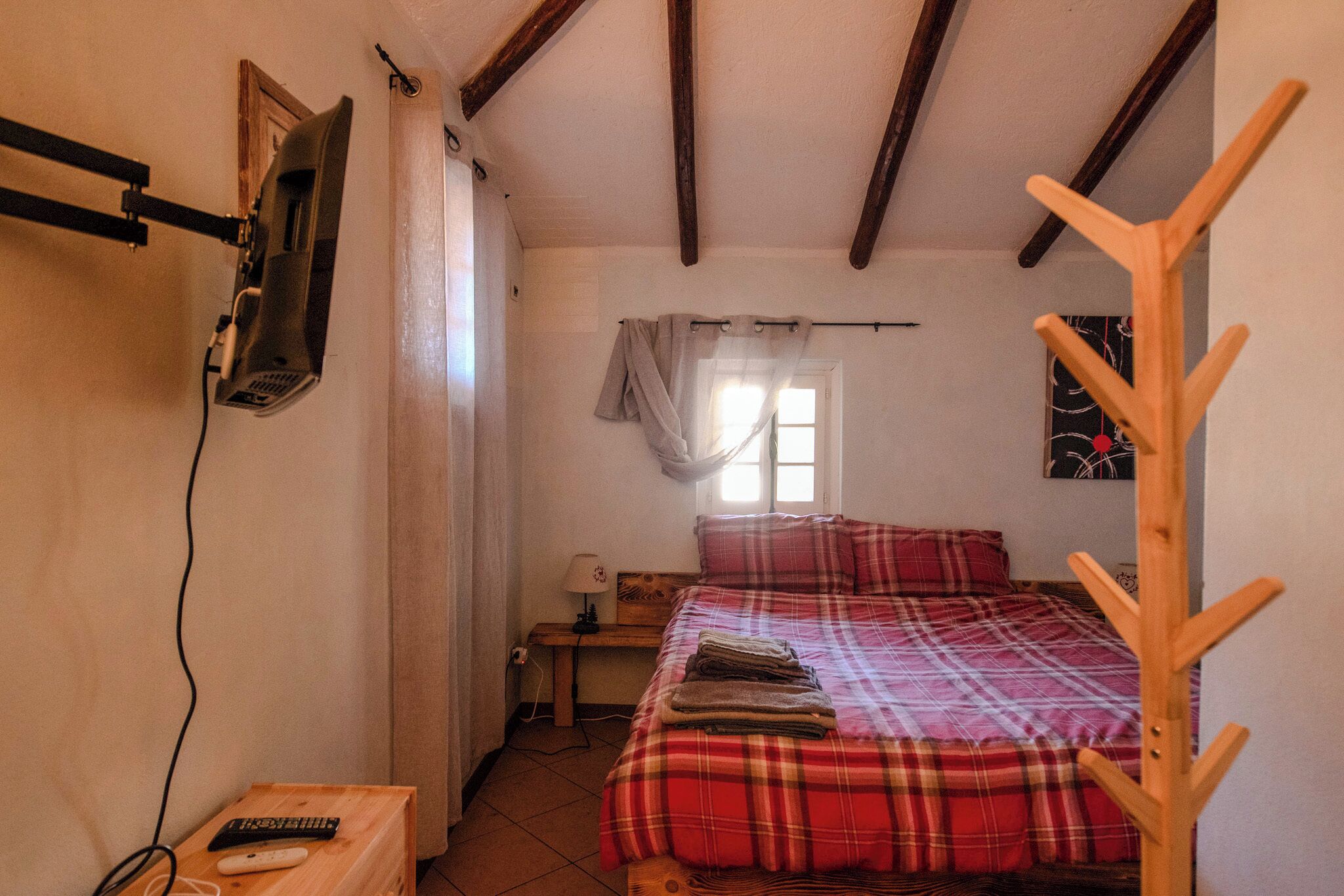 Un dormitorio con una cama, un banco de madera, un televisor montado en la pared y una ventana con cortinas.