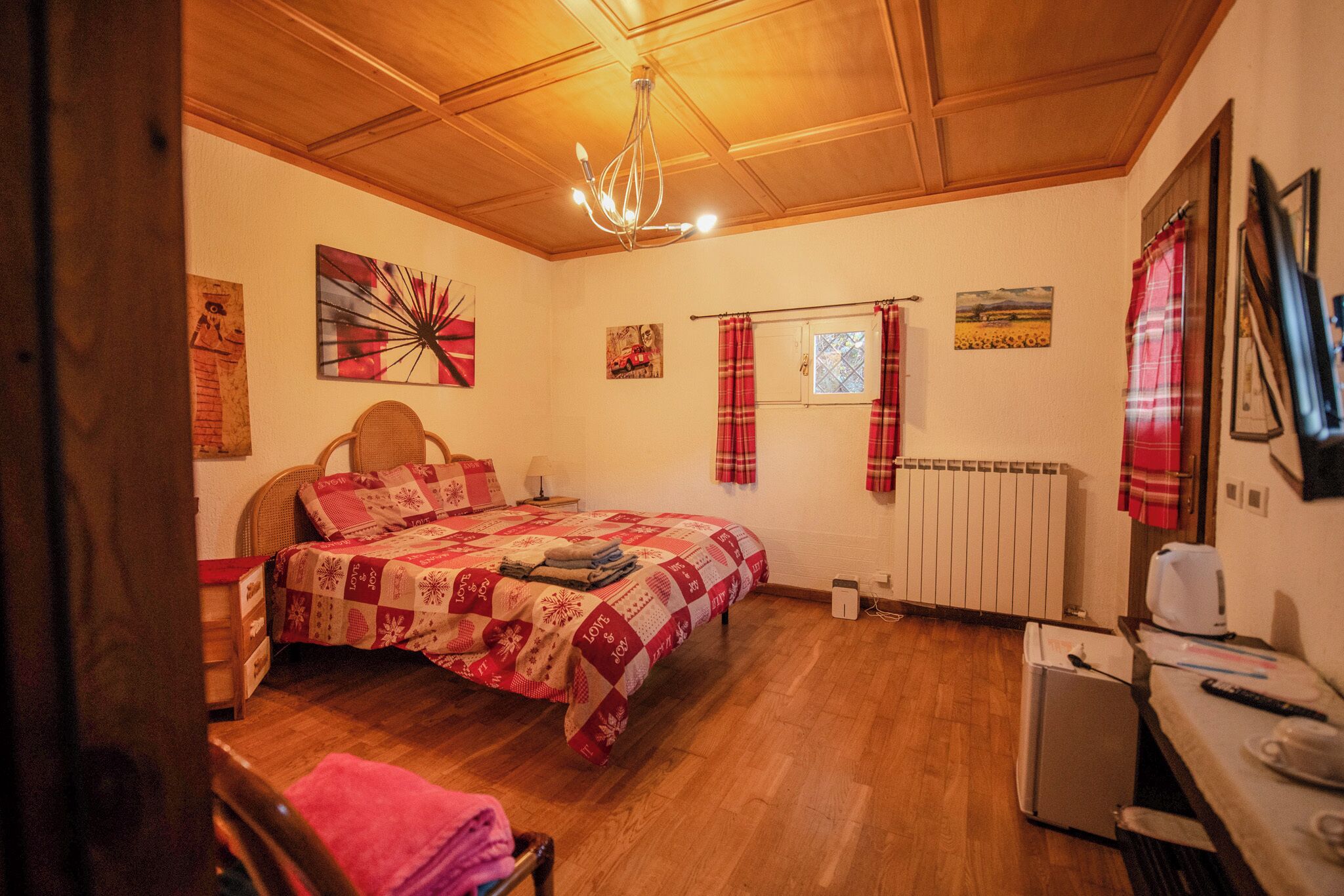 Un dormitorio acogedor con techo de madera, una cama con colcha roja y blanca, una mesita con hervidor y un radiador.