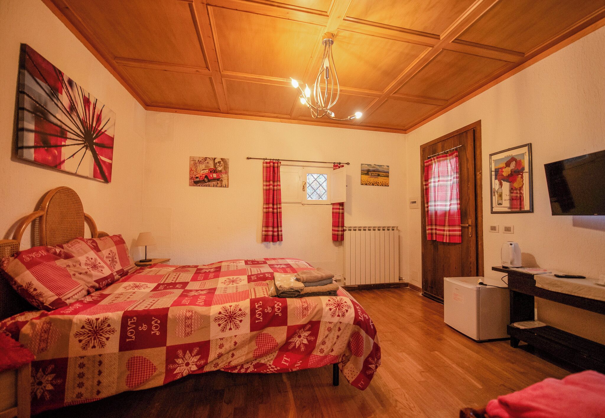 A cozy bedroom with a wooden ceiling, a bed with a red and white quilt, a small table with a lamp, and a radiator.