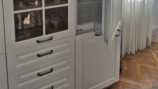 Un meuble blanc, doté de nombreux tiroirs et de portes en verre, contenant des ustensiles de cuisine et une bouteille.