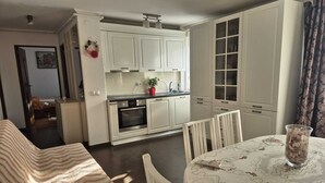 Une cuisine compacte avec des meubles blancs, une table à manger recouverte d’une nappe à motifs et une petite plante en pot.
