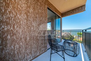 Un balcón con dos sillas y una mesa, un muro con textura y una vista despejada del cielo y del paisaje.