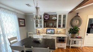Un comedor con una mesa de madera oscura y sillas blancas, una araña y una cocina con armarios blancos y salpicadero de ladrillo.