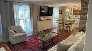 Sala de estar con sofá, sillón y mesa de centro. Hay un televisor montado en la pared, un comedor con mesa y sillas, y una zona de cocina con pared de ladrillo y un reloj.