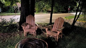 Dos sillas Adirondack y un pozo de fuego en un área cubierta de césped, con árboles y una carretera al fondo.