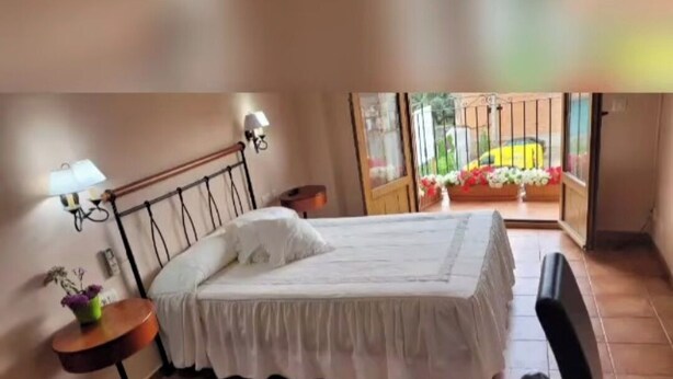 A bedroom with a bed, a small table with a lamp, and a view of a balcony with flowers. - Hotel Rural Cristania (Caminomorisco)