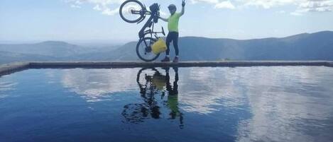 Una persona de pie sobre una superficie reflectante con una bicicleta, rodeada por un paisaje montañoso.