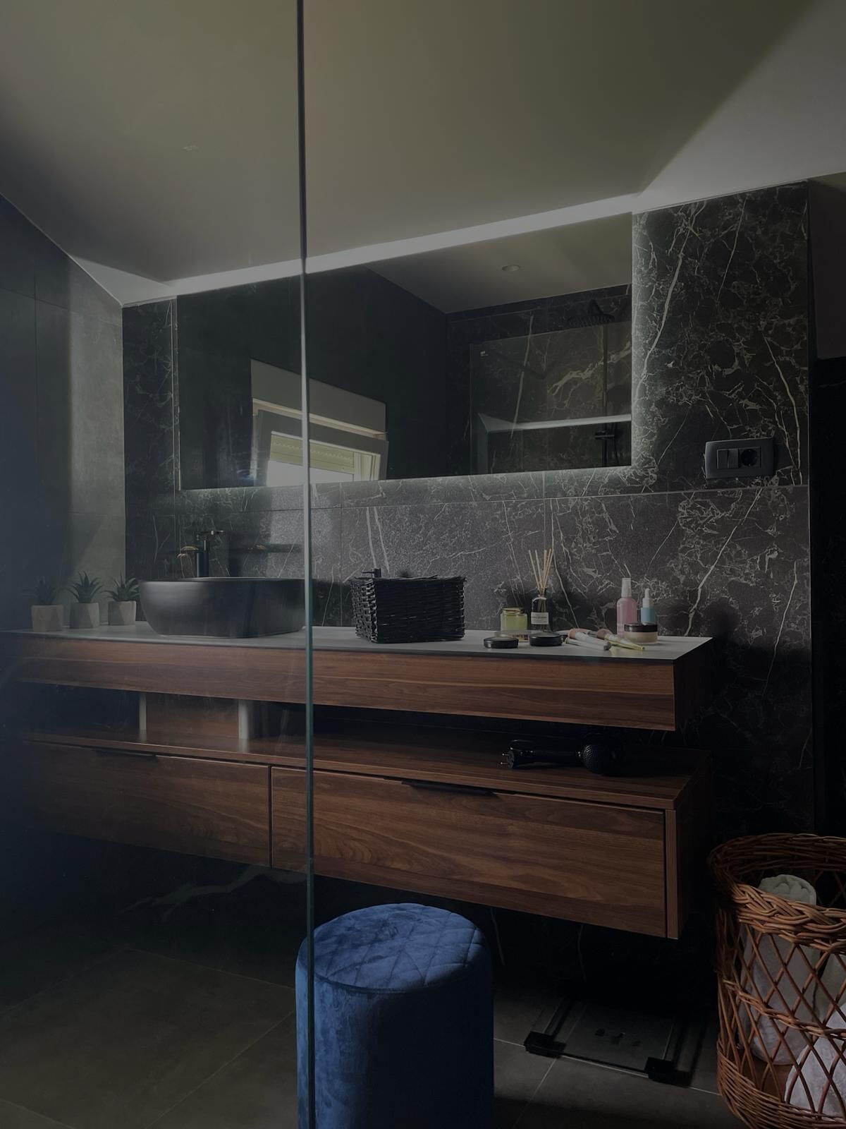 Une salle de bain moderne avec un meuble-lavabo en marbre foncé, un grand miroir et un pouf bleu.