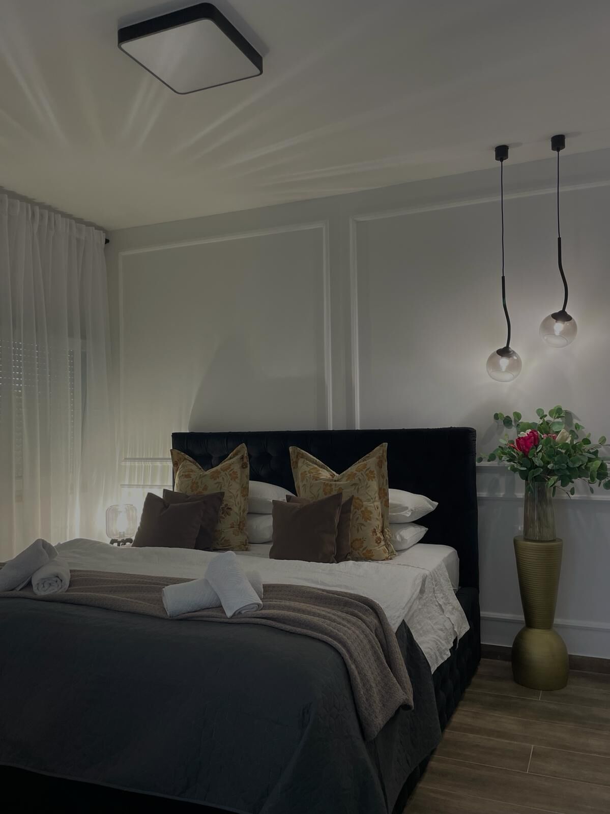 Una cama bien hecha con un cabecero oscuro, varios almohadones y un jarrón decorativo con flores.