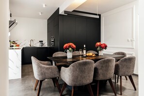Un espace repas moderne avec une table en bois foncé, six chaises grises à revêtement rembourré et, en arrière-plan, une cuisine noire.