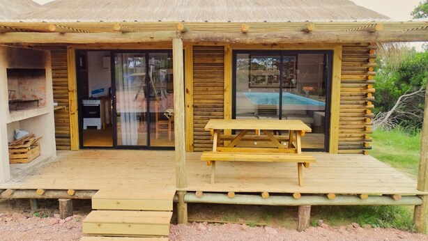 Uma cabana de madeira com telhado de palha, grandes portas de vidro deslizantes e um deck de madeira com mesa para piquenique.